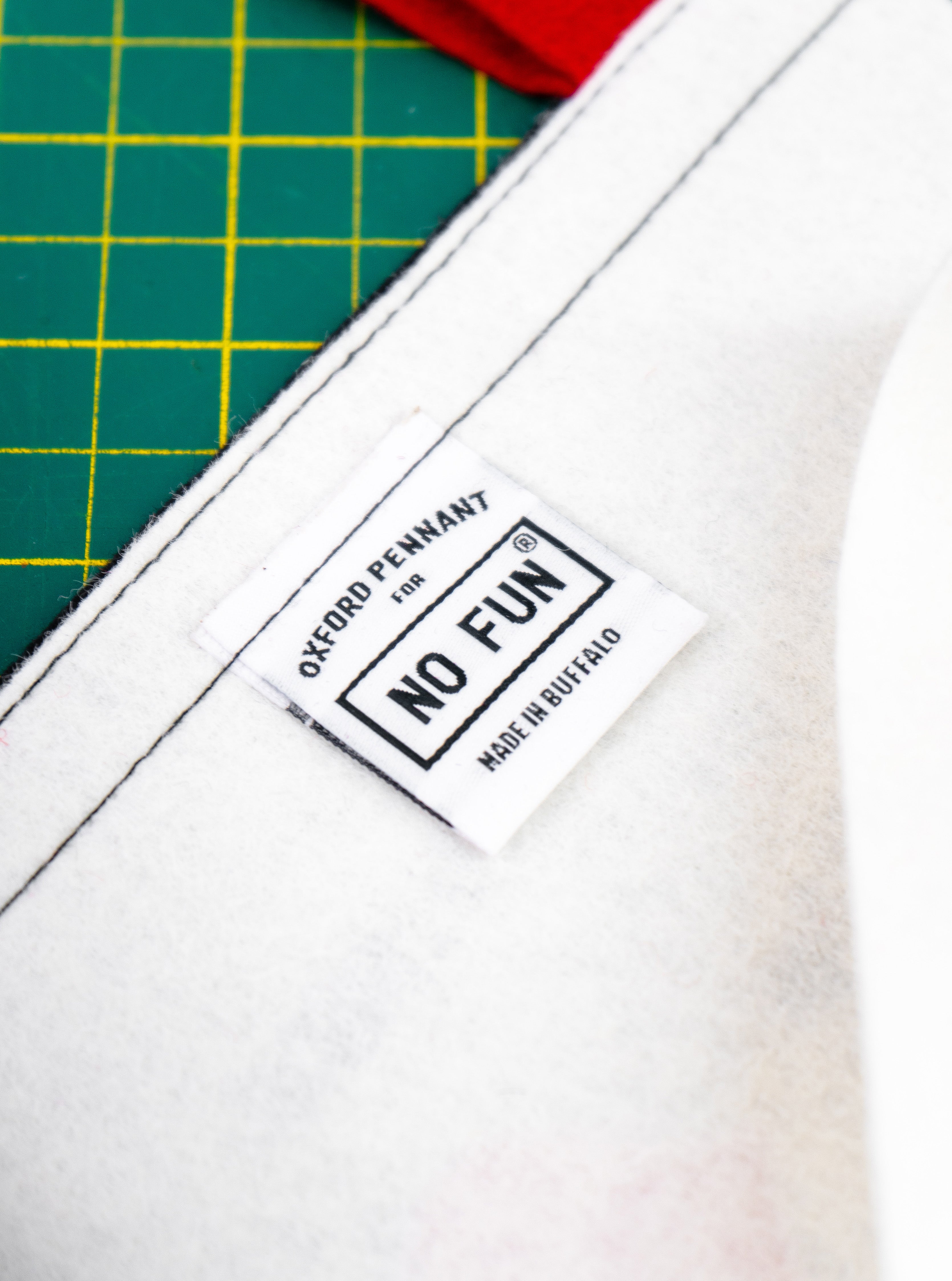 A white pennant with white woven tag that reads "Oxford Pennant for No Fun® made in Buffalo".  There is a green cutting mat in the background, and the edge of the red ties can also be seen.