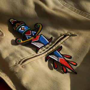 "Dagger" two-part embroidered iron-on patch designed by Brandon Ing. Patch is of traditional tattoo flash drawing of a dagger, split into two halves. One half is the blade, and the other half is the handle. Patch is multi colored, photographed against a tan, canvas fabric with a pocket paced between the split halves.