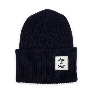 Photo of the "Life is Hell" Beanie by No Fun®.  Product is photographed against a white background.  Beanie is navy blue, and features a small white woven label on the cuff that reads "Life Is Hell".  Label is white, with black text, and red contrasting stitching.