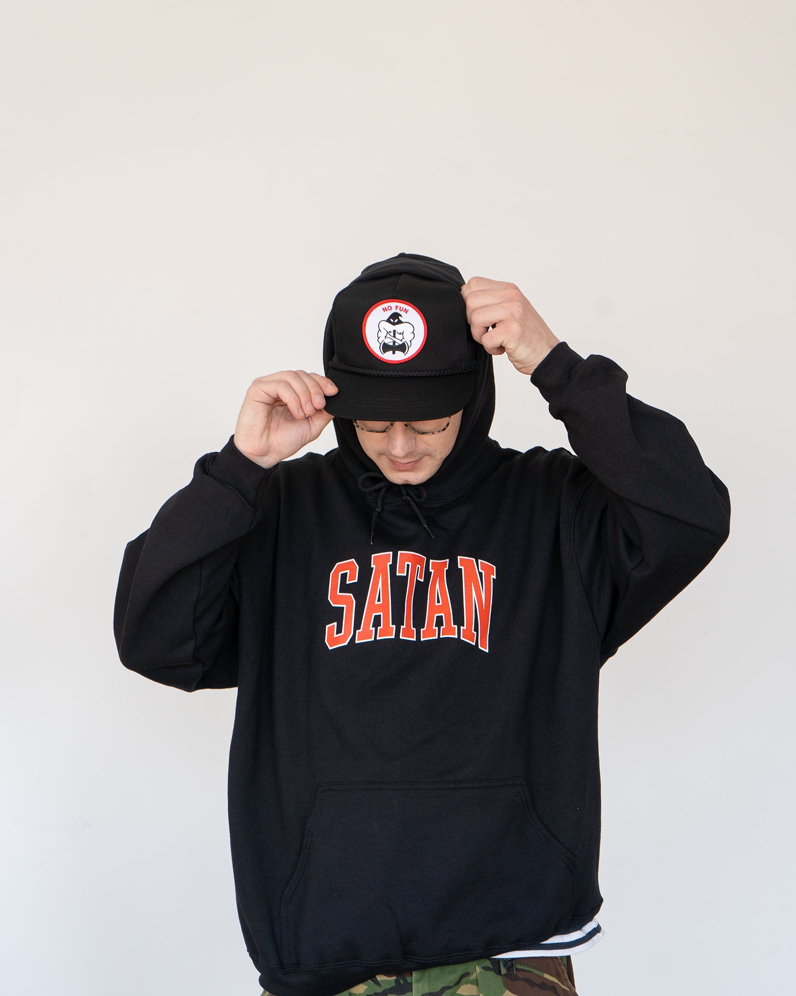 Photo of the "NO FUN®" X "Crawling Death" collaboration black, strapback hat.  The hat features a white patch with red embroidered border.  The embroidered graphic on the patch is a black and white executioner, with red "NO FUN®" logo.  This photo features a model in a hoodie wearing the hat.