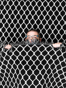 A person underneath the "Chainlink" woven blanket by No Fun®.  The person is peaking over the top of the blanket to showcase the scale of the design.  Only the top half of their face, and hands, are visible.