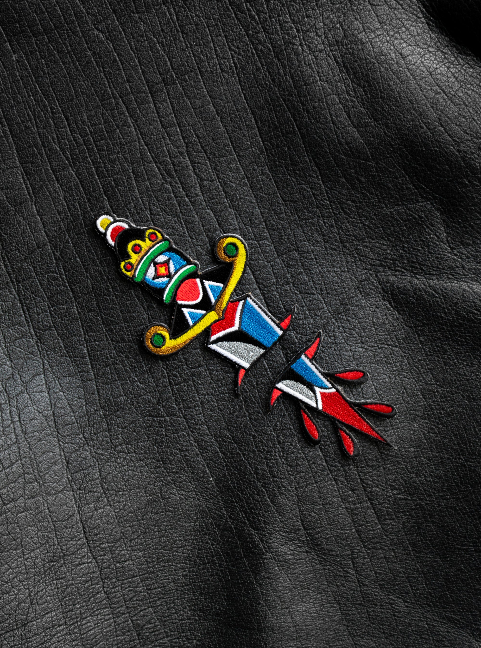 "Dagger" two-part embroidered iron-on patch designed by Brandon Ing. Patch is of traditional tattoo flash drawing of a dagger, split into two halves. One half is the blade, and the other half is the handle. Patch is multi colored, photographed against a leather jacket texture.