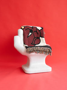 The "Devil" Woven Blanket folded, and draped over the back of a chair that is in the shape of a hand.  They are placed in front of a red background.