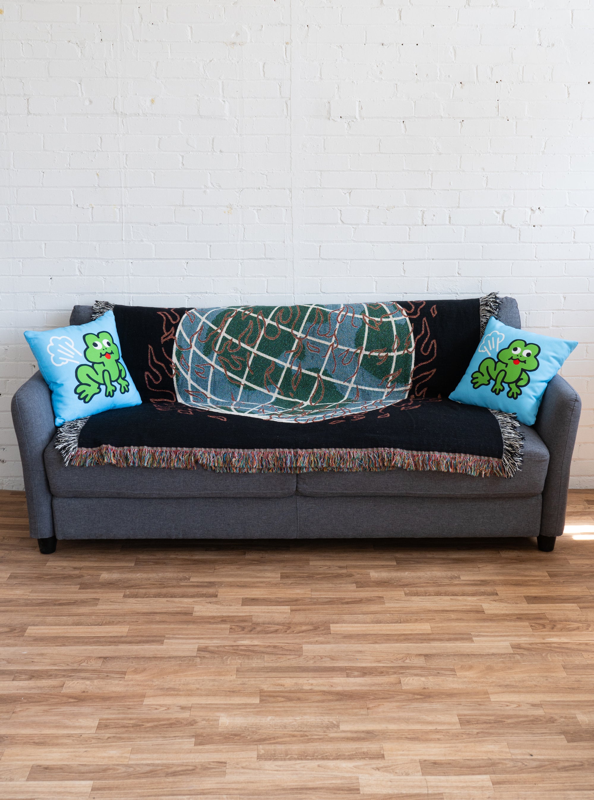 Two frog pillows displayed on the ends of a couch.  The couch is in a white room, with hardwood flooring.  The couch is grey, and has a woven blanket draped against the back of the couch.